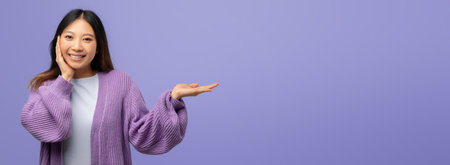 Smiling Young Woman in Purple Attire Gesturing Towards Empty Space on a Pastel Purple Backgroundの写真素材