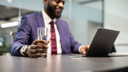 Positive black manager drinking water while working on laptop, panorama