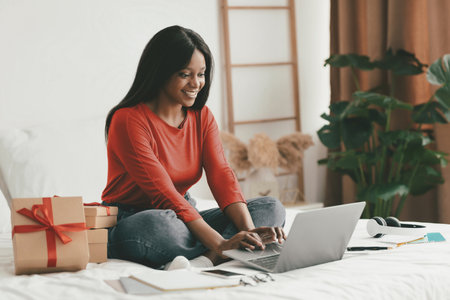 Smiling Woman Sitting on Bed With Laptop and Gifts in Cozy Room During the Dayの写真素材
