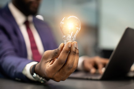 Black businessman holding illuminated light bulb, sitting in officeの写真素材