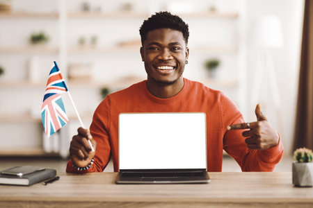 Smiling Young Man Holding a British Flag While Pointing at a Blank Laptop Screen Indoorsの写真素材