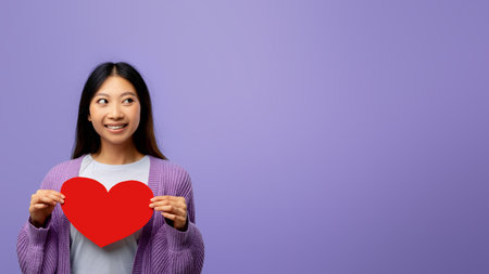 Woman Holding a Red Heart Shape Prop With a Purple Background During a Cheerful Momentの写真素材