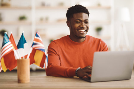 Man Smiling While Using Laptop in a Cozy Workspace With International Flags Aroundの写真素材