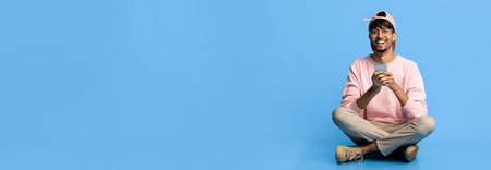 Young Man Sitting on Floor With Smartphone and Smiling Against Blue Background in Studio Settingの写真素材