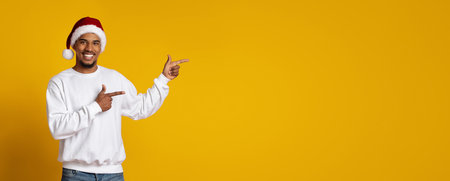 Cheerful Man Wearing Santa Hat Points to Empty Space Against Yellow Backgroundの写真素材