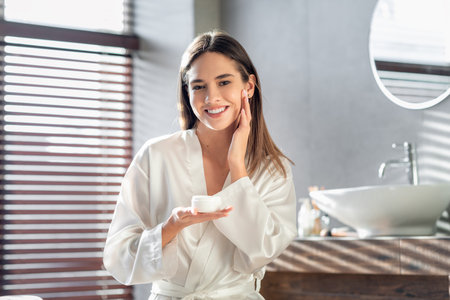 Young Woman Enjoys Skin Care Routine While Applying Moisturizer in Bright Bathroomの写真素材