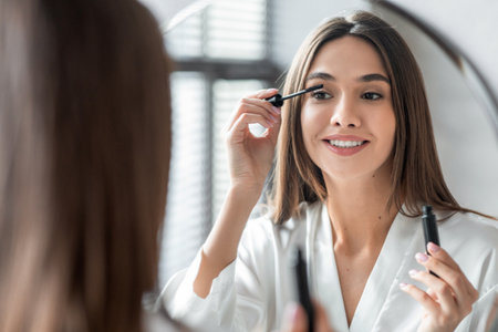 Lovely Young Woman Enhances Her Eyes by Applying Mascara in the Bathroom Mirrorの写真素材
