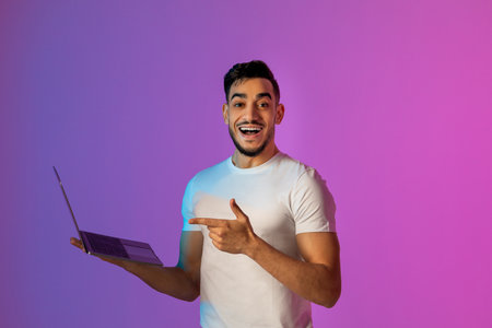 Cheery Young Arab Man Engaged in Online Work With Laptop in Vibrant Neon Lightの写真素材