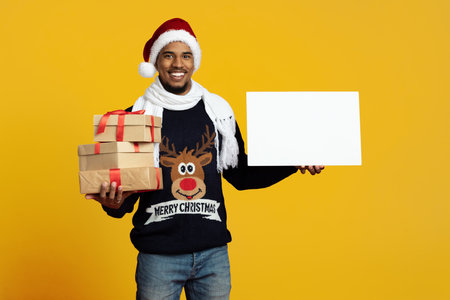 Cheerful Man in Christmas Sweater Holding Gifts and Blank Sign Against Yellow Backgroundの写真素材