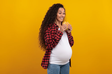 Excited Pregnant Woman Enjoying a Big Hamburger in a Bright Studio Settingの写真素材
