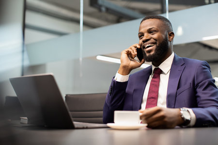 Positive african american businessman working on laptop, having phone conversationの写真素材