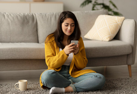 Positive Young Arab Woman Enjoys Coffee While Reading News on Smartphone in Cozy Living Roomの写真素材