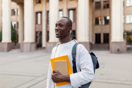 Student Smiling in Front of University Building During Daytime, Holding Books and Wearing a Backpackの写真素材
