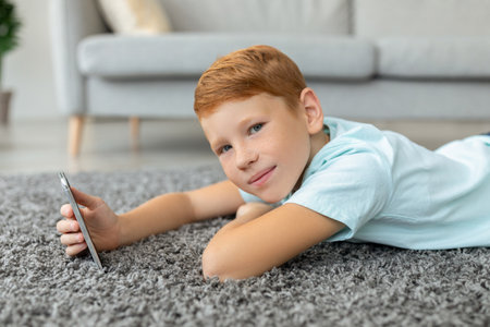 Redhead Boy Relaxing on Floor With Mobile Phone, Enjoying His New Gadget in Home Settingの写真素材