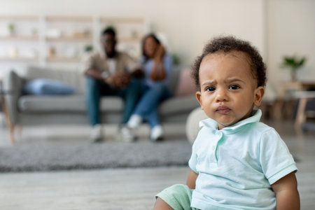Serious Angry African American Baby Boy Sitting on the Floor While Parents Relax on the Sofaの写真素材