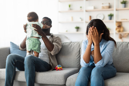 Exhausted African American Mom Struggles With Emotions While Dad Plays With Their Baby at Homeの写真素材