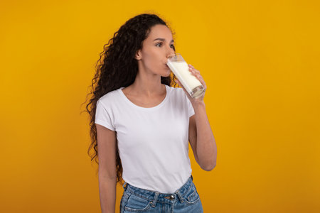 Happy Young Woman Enjoying a Glass of Milk in a Studio Setting With Yellow Backgroundの写真素材