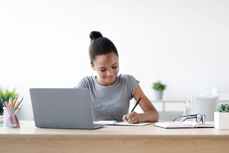 Young African American Girl Studies at Home, Writing Notes and Using a Laptop During COVID-19の写真素材