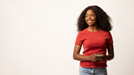 Beautiful Young Black Female Smiling While Holding Smartphone and Looking Aside With Confidenceの写真素材