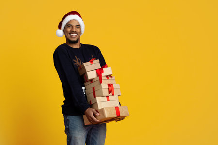 Smiling Man in Santa Hat Carries Stacked Holiday Presents Against Bright Yellow Backgroundの写真素材
