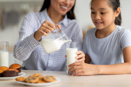Asian Mother Pours Fresh Milk for Daughter During Breakfast in the Home Kitchenの写真素材