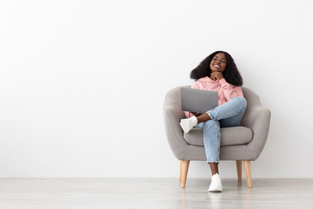 Smiling Woman Sitting in a Cozy Chair While Using a Laptop in a Bright, Modern Roomの写真素材