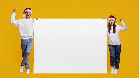 Cheerful Couple in Santa Hats Holding Blank Sign Against Bright Yellow Backgroundの写真素材