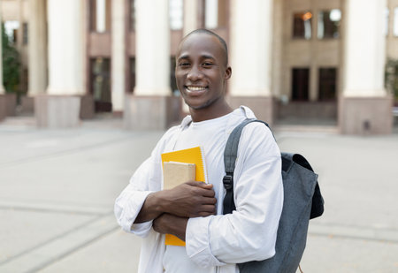 Smiling Student Holding Books Stands Confidently on Campus in Sunny Weatherの写真素材