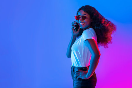 Woman in Stylish Sunglasses Smiles While Talking on Her Phone in Colorful Lightingの写真素材