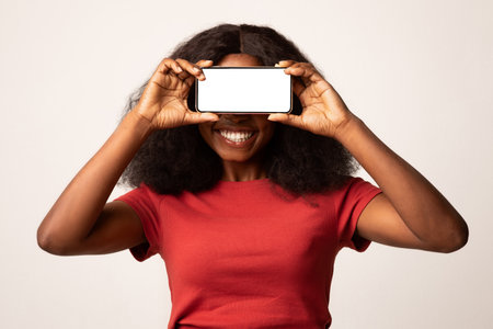 Cheerful Black Woman Covering Eyes With Blank Smartphone in a Bright Studio Settingの写真素材