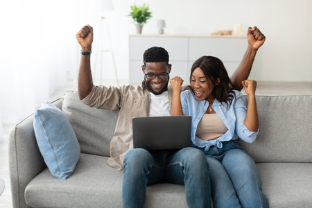 Excited Couple Celebrates Victory While Using Laptop at Home on a Cozy Couchの写真素材