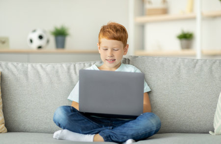 Redhead Child Enjoys Gaming on Laptop Alone at Home in Modern Living Room Settingの写真素材