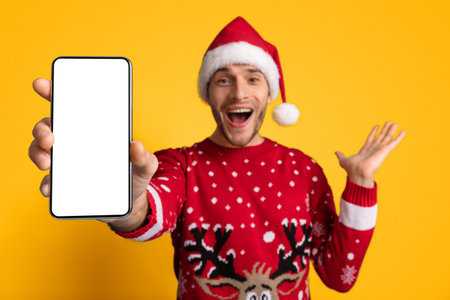 Joyful Man in a Christmas Sweater Showing a Smartphone Against a Bright Yellow Backgroundの写真素材