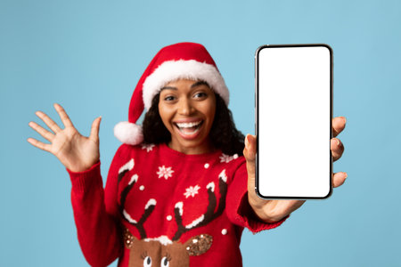 Woman in Christmas Sweater Smiles and Waves While Holding a Blank Smartphone Against a Light Blue Backgroundの写真素材