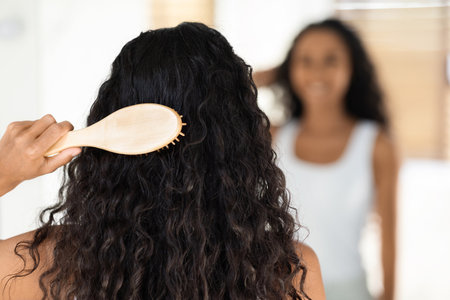 Young Woman Brushes Her Curly Hair While Looking at Her Reflection in Bright Roomの写真素材