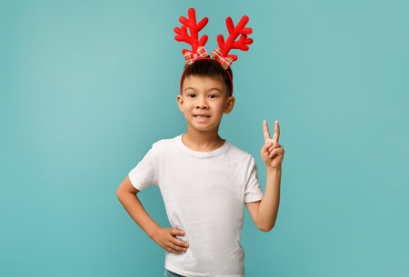 Child With Red Reindeer Antlers Smiling and Showing Peace Sign Against a Blue Background During Festive Seasonの写真素材