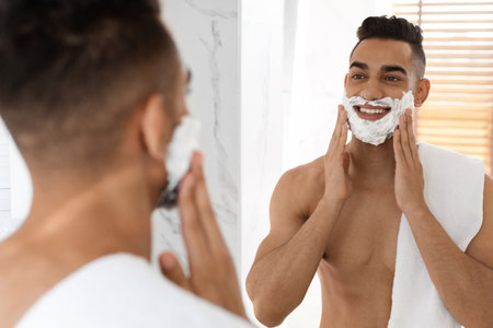 Man Enjoying a Morning Shave While Smiling in a Bright Bathroom With a Towel Around His Shoulderの写真素材