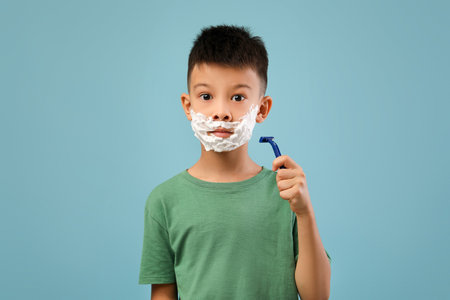Young Boy With Shaving Cream on Face Holding a Razor, Showcasing Early Grooming Skills Against a Blue Backgroundの写真素材