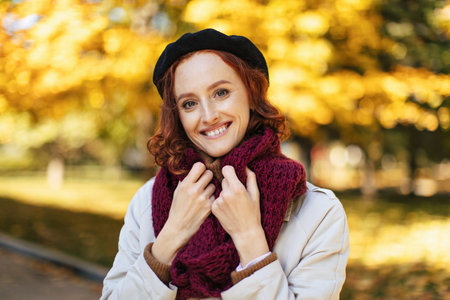 Woman in Stylish Beret Smiling Warmly While Posing Outdoors Surrounded by Autumn Leaves and Trees in Sunny Park Settingの写真素材