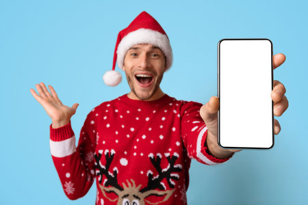 Man in Festive Sweater and Santa Hat Celebrates With Smartphone Against a Blue Background During Holiday Seasonの写真素材