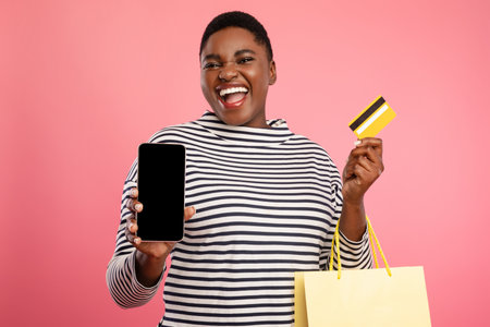 Happy Shopper Showing Mobile Phone and Credit Card in Front of Pink Background While Holding...