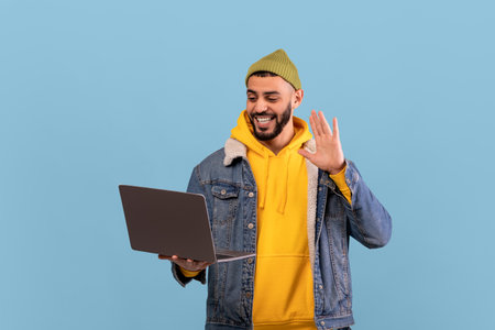 Young Man in a Yellow Hoodie Waves While Using a Laptop Against a Blue Background