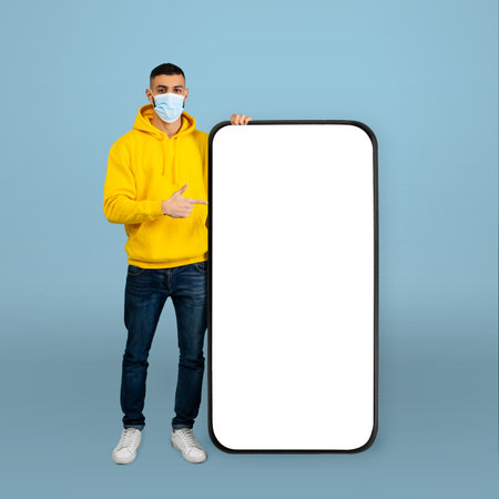 Young Man in Yellow Hoodie Points to Blank Board in Studio With Blue Background While Wearing a Mask