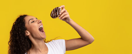 Woman Happily Preparing to Eat a Chocolate Donut Against a Bright Yellow Backgroundの写真素材