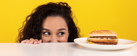 Excited Woman Peeking at a Delicious Burger on a Plate Against a Bright Yellow Backgroundの写真素材