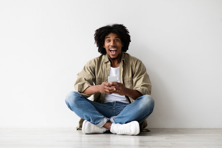 Excited Black Man Reacts Joyfully to News While Sitting on the Floor With Smartphoneの写真素材