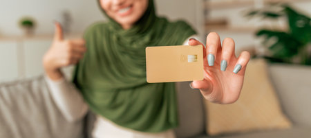 Woman Showing a Contactless Card While Smiling and Giving a Thumbs up in a Cozy Living Roomの写真素材