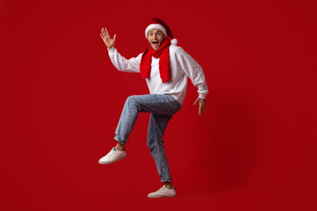 Man Wearing Santa Hat and Red Scarf Happily Dancing Against a Bright Red Background During Festive Seasonの写真素材