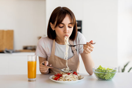 Young Woman Enjoying a Delicious Pasta Meal With Fresh Vegetables and Juice at Home on a Bright Afternoonの写真素材