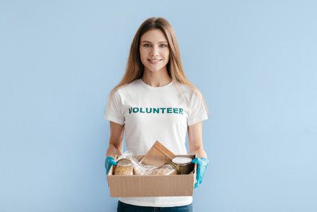 Smiling Volunteer Holds Care Package With Food Items Against a Light Blue Backgroundの写真素材